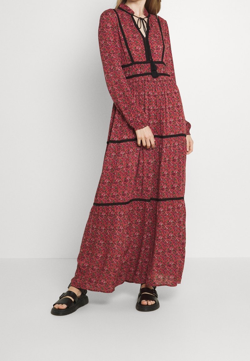 Femme portant une longue robe rouge à fleurs avec un biais noir et des sandales noires, debout devant un fond blanc uni.
