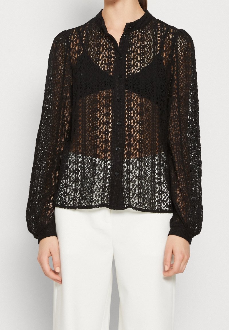 Blouse en dentelle noire à manches longues, boutonnée sur le devant avec un col montant, portée sur un soutien-gorge noir, associée à un pantalon blanc.