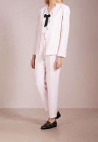 Vaaleanpunainen räätälöity puku, jossa on aaltoileva bleiseri ja kerma-valkoinen paita, jossa on musta solmuke. Mustat kengät, joissa on niittikoristeita, täydentävät asun.