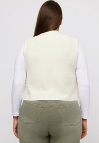 Cremefarbene Strickweste mit strukturierter Oberfläche, kurzem Schnitt und Rundhalsausschnitt, kombiniert mit olivgrünen Hosen mit Gesäßtaschen.