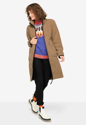 Bruine fleece jas met een hoge kraag, gecombineerd met een blauw gebreid sweater met geometrische accenten en zwarte broek. Dikke witte laarzen.