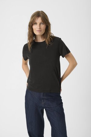 Femme aux cheveux châtain clair portant un t-shirt noir uni à manches courtes et un jean bleu foncé, debout avec les mains derrière le dos.