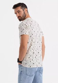 Kortærmet t-shirt i beige med sort vertikalt tekstmønster; har rund halsudskæring og en afslappet pasform, parret med lys blå jeans.