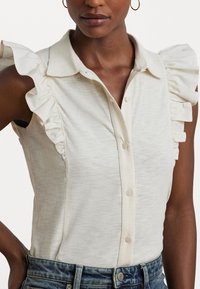 Blouse sans manches beige clair avec des accents de volants sur les épaules, le col et le devant à boutons ; tissu texturé et design ajusté.