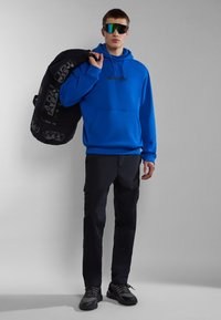Sudadera azul con bolsillo frontal y logo, pantalones cargo negros, mochila negra con gráficos y gafas de sol con lentes reflectantes verdes.