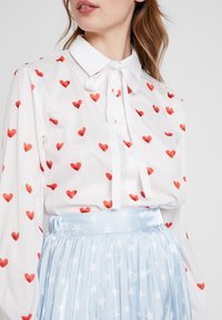 Vit blus med spridda röda hjärttryck, med krage och detaljer i form av fluga. Kombinerad med en ljusblå plisserad kjol prydd med vita stjärnor.