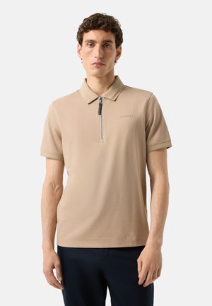 Mand iført en beige kortærmet polo med frontlynlås og mørke bukser, stående mod en ensfarvet lys baggrund.
