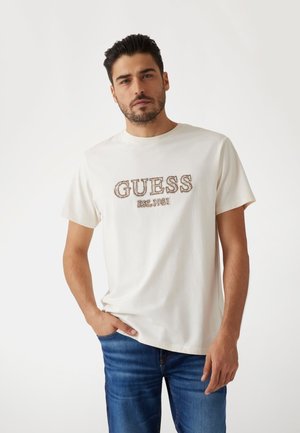Krem majica od pamuka s "GUESS EST.1981" logotipom u smešnoj koncu, okrugli izrez, kratki rukavi i opušteni kroj, upotpunjena plavim farmerkama.