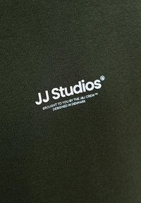 Tkanina w kolorze oliwkowym o drobnej fakturze, z białym napisem: "JJ Studios" oraz hasłem poniżej, wyśrodkowane i wyraźne, na swobodnej odzieży.