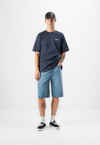 Navy bomulls t-skjorte med grafikk, lys blå denimshorts, grå sokker og svarte sneakers. Modell iført en svart caps, står mot en hvit bakgrunn.