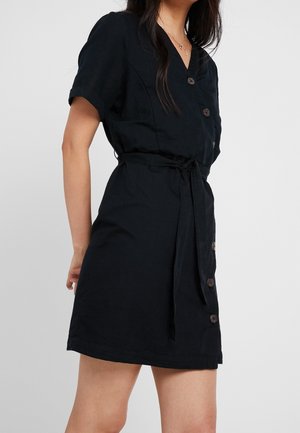 Kobieta ubrana w czarną sukienkę na guziki z krótkim rękawem, zawiązaną paskiem w talii oraz warstwowymi złotymi naszyjnikami, stoi na tle szarego tła.