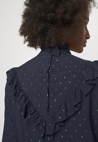Personne aux cheveux bouclés portant un chemisier bleu foncé à pois argentés, avec des boutons et des détails froncés dans le dos et aux épaules.