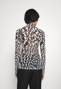 Persona con capelli neri corti che indossa un top a maniche lunghe e collo alto con un motivo astratto ondulato in bianco e nero, in piedi su uno sfondo bianco.