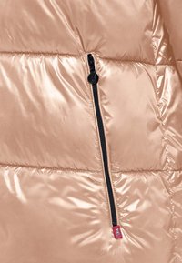 Sekcja różowej złotej kurtki puchowej z błyszczącą, pikowaną teksturą. Posiada czarny zamek błyskawiczny oraz małą czerwoną metkę z logo w pobliżu zamka.