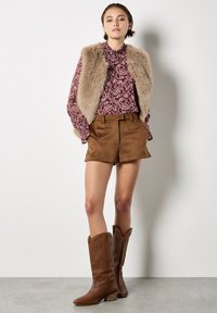 Pantaloni corti in suede marrone, blusa in bordeaux a fantasia e gilet in pelliccia sintetica. Completato con stivali da cowboy in pelle marrone con cuciture dettagliate.