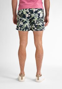 I pantaloni da bagno da uomo presentano uno sfondo blu navy con un motivo di foglie e fiori tropicali in verde e bianco. Abbinati a sneaker bianche.
