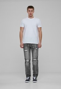 Maglietta bianca a maniche corte abbinata a jeans grigi strappati con ginocchia sfilacciate. Il modello indossa sneakers nere con suole bianche.
