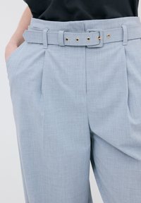 Lichtblauwe broek van een gestructureerde stof, met een tailleband met gouden hardware en scherpe plooien aan de voorkant.