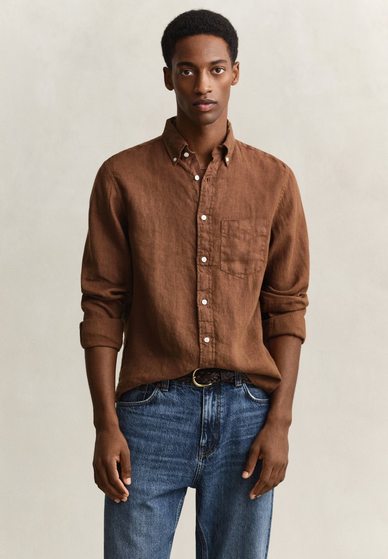 Jeune homme portant une chemise marron à manches retroussées et un jean bleu, debout devant un fond beige uni, faisant face à l'avant.