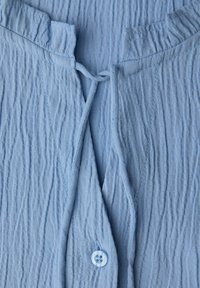 Světle modrá mačkaná halenka s předním zavazováním, zdobená knoflíkem a texturovanou látkou, která přidává rozměr.