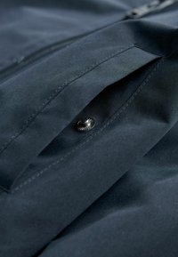 Temno modra tkanina z gladko teksturo, s vidnim žepom in dekorativnim srebrnim gumbom na pritrditev. Čist, minimalističen dizajn.