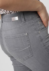 Des jeans en denim gris présentent une taille haute, une coupe slim et des poches arrière avec des coutures décoratives. La ceinture comporte une étiquette en métal avec un logo.