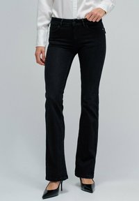 Mujer con jeans bootcut negros, camiseta blanca de manga larga y tacones altos negros de punta, de pie contra un fondo liso.