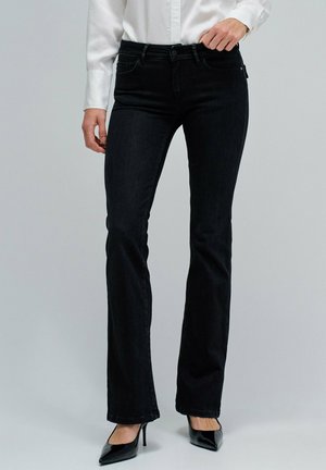 Mujer con jeans bootcut negros, camiseta blanca de manga larga y tacones altos negros de punta, de pie contra un fondo liso.