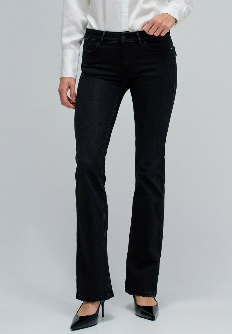 Mujer con jeans bootcut negros, camiseta blanca de manga larga y tacones altos negros de punta, de pie contra un fondo liso.