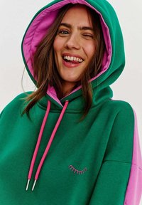 Zelená mikina s ružovým vnútorným podšívkou, ružovými šnúrkami a grafikou rias. Má pohodlný strih a mäkkú textúru.