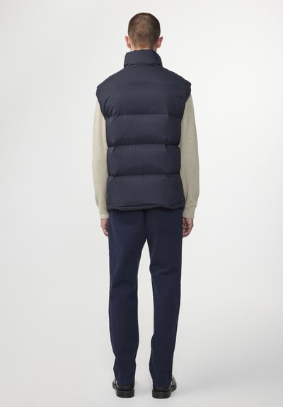 Marineblau weste ohne Ärmel mit hohem Kragen, ausgestattet mit einem gesteppten Design. Kombiniert mit einem cremefarbenen Pullover und dunklen Hosen.