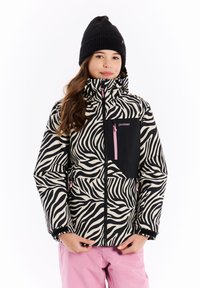 Nuori henkilö, jolla on päässään musta pipo, zebra-kuvioinen takki, jossa on vaaleanpunaiset vetoketjut, ja vaaleanpunaiset housut, seisoo valkoista taustaa vasten.