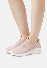 Sapatilhas rosa claro, tipo slip-on, com um superior em malha texturizada, sola branca e detalhes contrastantes no calcanhar. Fechadas, sem atacadores visíveis.