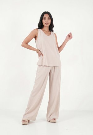 Femme debout, main sur la hanche, portant un haut beige sans manches ample, un pantalon large, des sandales à plateforme et un bracelet sur fond blanc.