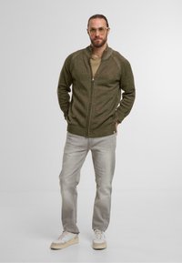 Olive green zip-up sweater with textured fabric, slim fit. Paired with light gray jeans and beige/white sneakers. Clean, modern design.