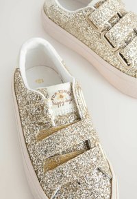 Baskets dorées pailletées avec une surface texturée, dotées de deux bandes Velcro, d'un intérieur blanc et d'une étiquette de marque indiquant "Jours de plaisir et de bonheur."