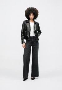 Koharate juustega naine, kes kannab musta nahktagi, valget särk, laiade säärtega musti teksapükse ja musti terava ninaga kontsakingi, seisab valge tausta ees.