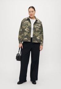Veste imprimée camouflage dans des tons de vert, dotée de deux poches poitrine et d'une fermeture à bouton, associée à un pantalon large noir et un petit sac noir.