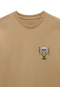 Beige puuvillapaita, jossa on graafinen kuva pääkallosta maljassa, ja sen vieressä on tekstit "VANS" ja "1966". Pyöreä kaula-aukko. Lyhyet hihat.