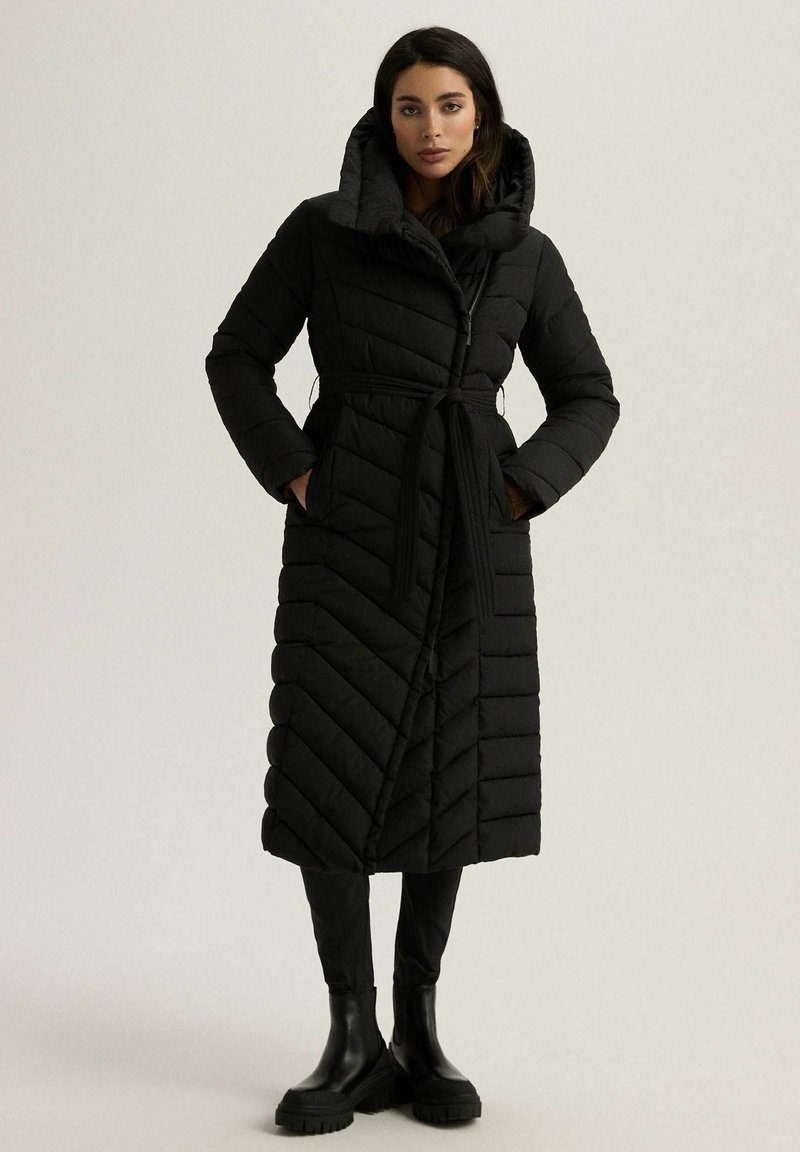 Długa czarna puchowa kurtka w wzór w zygzak, z wysokim kołnierzem i wiązanym paskiem w talii. Posiada boczne kieszenie i długie rękawy. Wykonana z materiału pikowanego.
