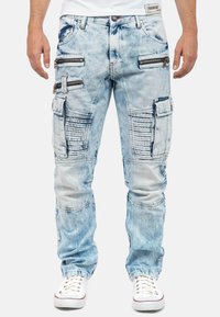 Light blue denim cargo pants with multiple zippered pockets, ribbed accents on legs, and a relaxed fit, paired with white sneakers.