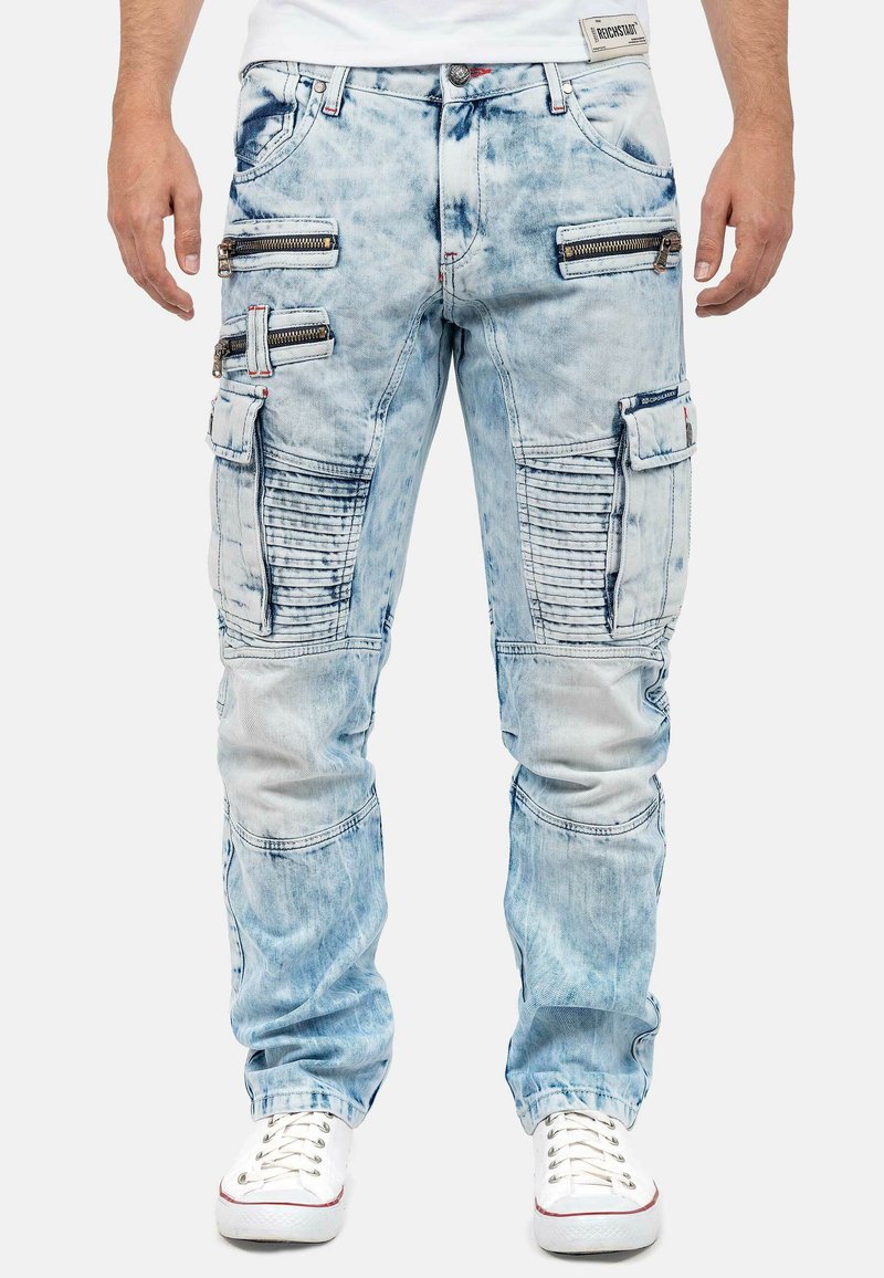 Light blue denim cargo pants with multiple zippered pockets, ribbed accents on legs, and a relaxed fit, paired with white sneakers.