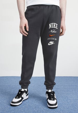 Person trägt schwarze Nike-Sweathosen mit mehreren Nike-Logos am linken Bein und schwarz-weiße Nike-Sneakers auf blauem Teppich.