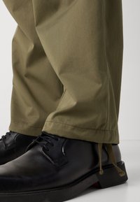 Olivgrüne Hosen mit glatter Textur, kombiniert mit schwarzen Lederschuhen mit Schnürsenkeln und einer strukturierten Sohle.