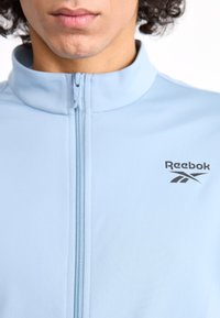 Light blue Reebok jacket with a high collar, front zipper, and a black logo on the left side. Smooth fabric texture and athletic design.