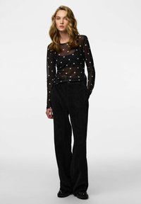 Czarna przezroczysta bluzka z długim rękawem w białe grochy, zestawiona z wysokim stanem czarnych sztruksowych spodni o szerokich nogawkach oraz błyszczące czarne buty.