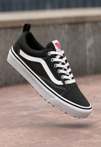 Juodai baltas Vans Old Skool sportbačiai su baltomis raišteliais, pakibę virš plytelių grindinio ir pilkos akmeninės sienos fone.