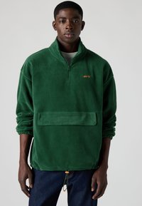 Zaļš flīsa pulovers ar puszipa apkakli, priekšējo kabatu un elastīgiem aproču galiem. Uz tā ir oranža Levi's logotips un aukla apakšmalā.