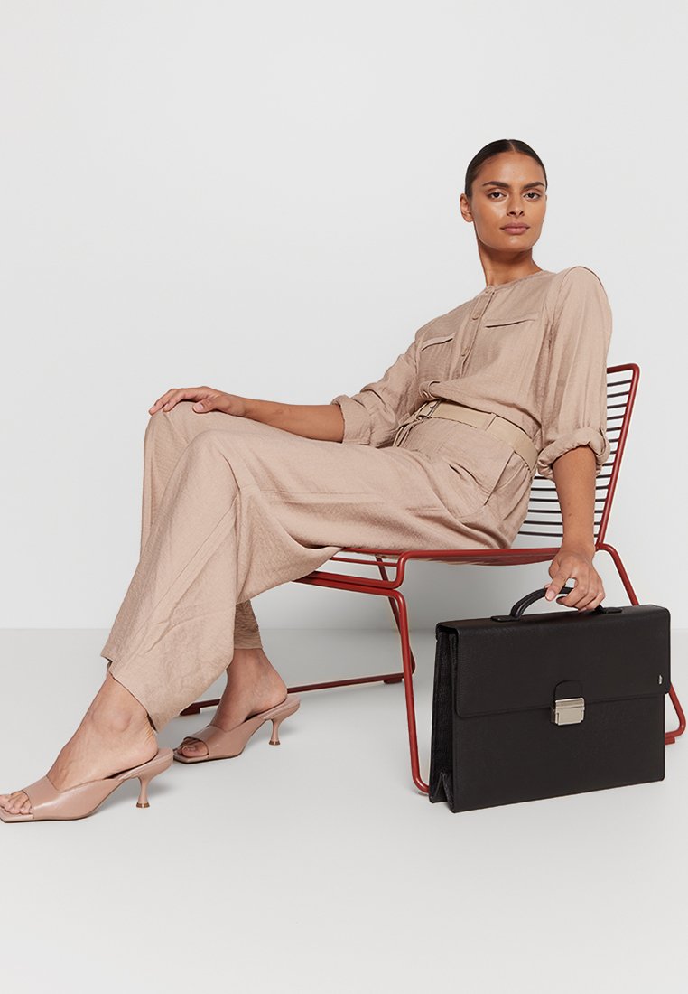 Beige jumpsuit met twee borstzakken en een ceintuur in de taille, gecombineerd met nude hoge muiltjes, terwijl er een zwarte leren aktetas met zilveren sluiting wordt vastgehouden.