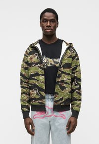 Φούτερ με φερμουάρ σε καμουφλάζ πράσινο και μαύρο, μείγμα βαμβακιού, διαθέτει κουκούλα με κορδόνι και ριμπ τελείωμα. Φορεμένο πάνω από μια μαύρη γραφική μπλούζα.